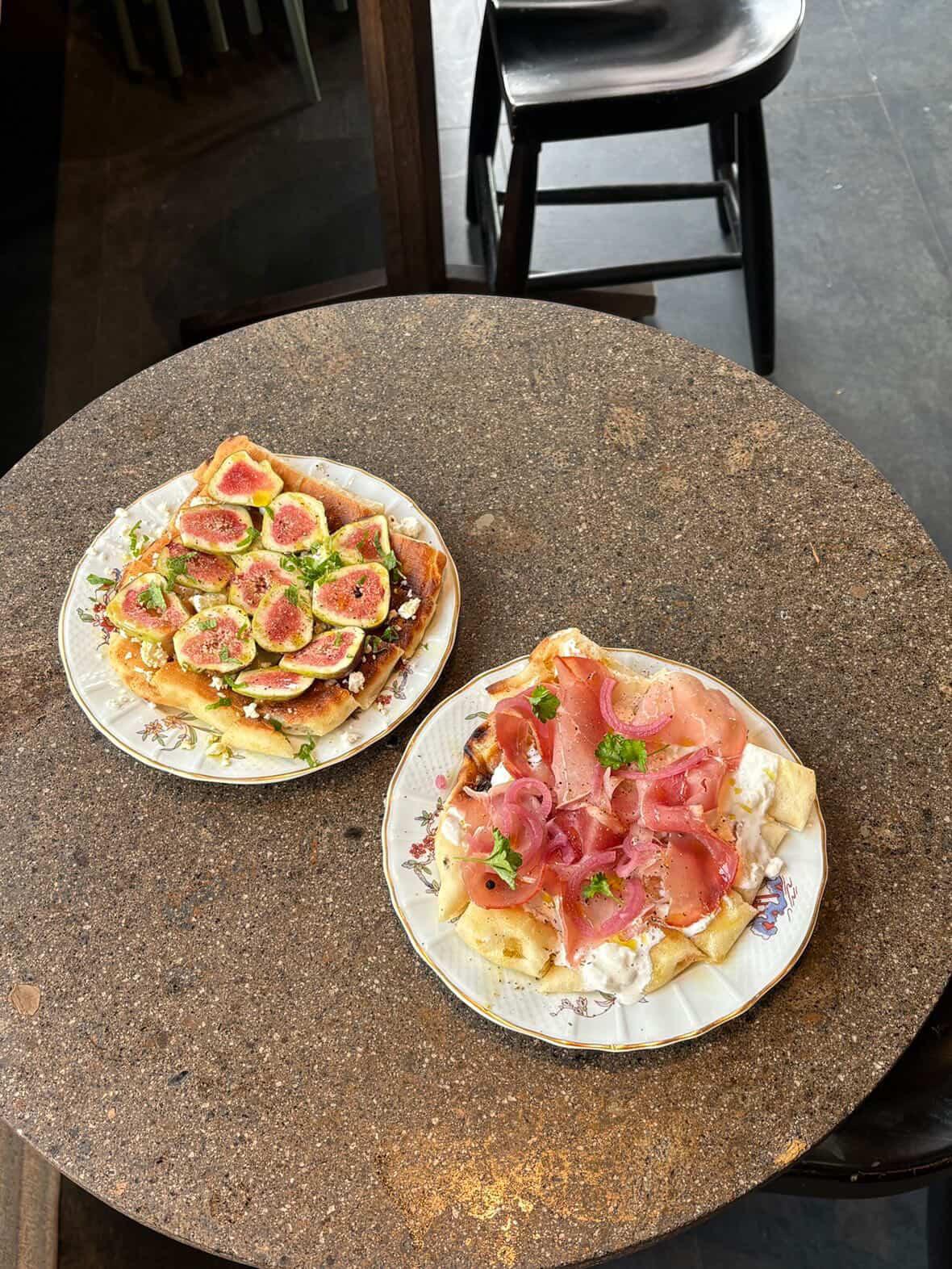 A fig flatbread and a coppa flatbread with burrata on a round stone table